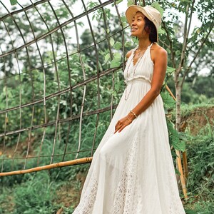 May include: A woman wearing a white halter dress with a lace hem and a straw hat, standing on a wooden bridge. The dress has a flowing silhouette. The background features a metal archway and lush greenery.
