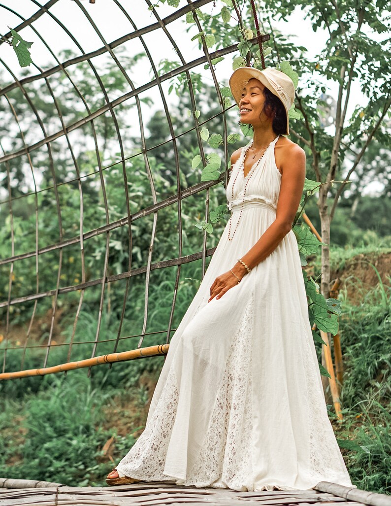 May include: A woman wearing a white halter dress with a lace hem and a straw hat, standing on a wooden bridge. The dress has a flowing silhouette. The background features a metal archway and lush greenery.