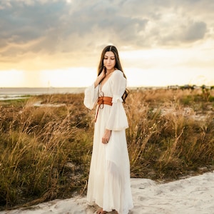 May include: A woman in a white maxi dress with a brown leather belt stands in a field of tall grass. The sky is cloudy and the sun is setting in the background.