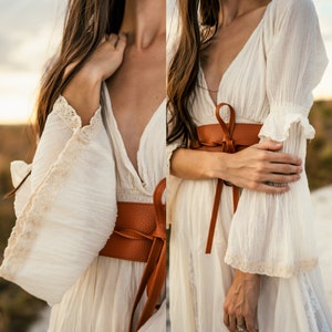 May include: A white, flowy dress with a deep V-neckline and lace trim on the sleeves. A brown leather belt is tied around the waist.
