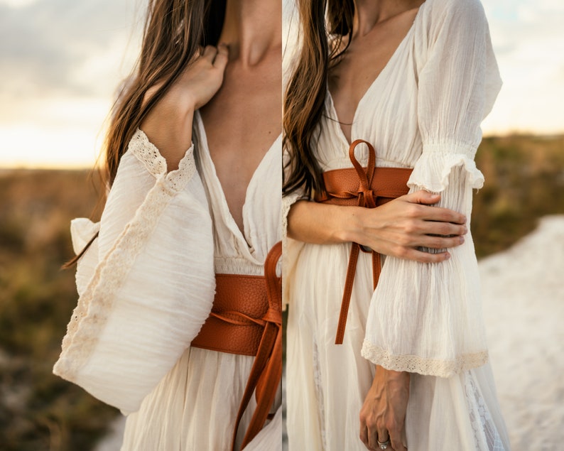 May include: A white, flowy dress with a deep V-neckline and lace trim on the sleeves. A brown leather belt is tied around the waist.