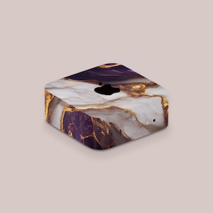 May include: A square-shaped electronic device with a marble design. The design features a blend of white, purple, and gold colors. The device has a black logo on the top and several small ports on the side.