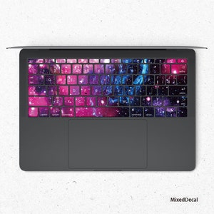 Könnte beinhalten: Eine Laptop-Tastatur mit einer Galaxie-bedruckten Haut. Die Tasten sind mit einem farbenfrohen Design aus rosa, blauen und violetten Nebeln mit weißen Sternen bedeckt.