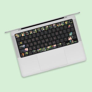 Könnte beinhalten: Eine Laptop-Tastatur mit floral gemusterten Aufklebern. Die Aufkleber zeigen eine Vielzahl bunter Blumen und Blätter auf schwarzem Hintergrund. Der Laptop ist silber und weiß, vor einem hellgrünen Hintergrund.