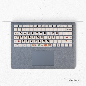 May include: A laptop keyboard with autumn-themed keycaps. The keys feature images of leaves, acorns, pumpkins, and squirrels. The keyboard is beige and has a gray trackpad.
