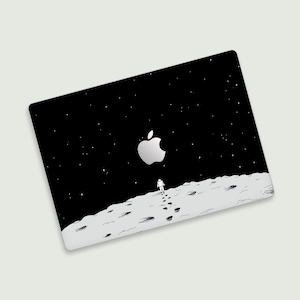 Op de afbeelding: Een zwarte laptop skin met een wit Apple-logo en een tekening van een astronaut die op de maan loopt met voetafdrukken.