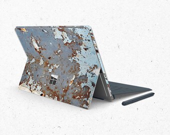 May include: A blue and brown laptop skin with a rusty, weathered design. The skin is applied to a closed laptop with a black keyboard and a black stylus.