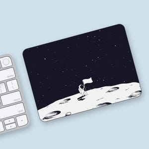 May include: A black and white illustration of an astronaut standing on the moon with a flag. The astronaut is wearing a spacesuit and holding a flag. The moon is depicted as a gray surface with craters. The background is a dark blue sky with stars.