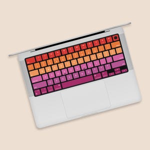 Puede incluir: Un ordenador portátil con una cubierta de teclado colorida. La cubierta del teclado tiene un degradado de rojo, naranja, amarillo y rosa. Las teclas están etiquetadas con el alfabeto estándar y los números.