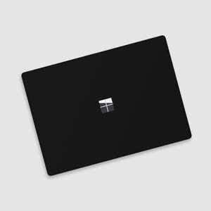 May include: A black laptop computer with the Microsoft Windows logo in the center. The laptop is closed and has a sleek, minimalist design, with a matte finish. The logo is a square made up of four smaller squares in shades of gray and white.