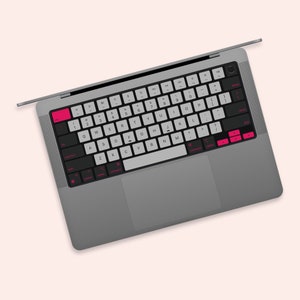 Puede incluir: Un ordenador portátil gris con un teclado negro y un trackpad. El teclado tiene teclas blancas con letras negras y acentos rosas. Las teclas están etiquetadas con letras, números y símbolos.