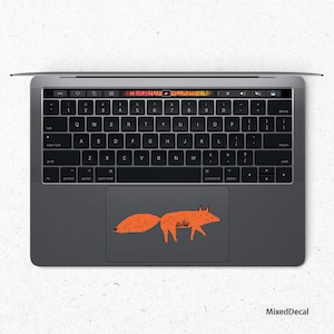 May include: A gray laptop computer with a orange fox illustration on the trackpad. The laptop has a standard keyboard with black keys and a silver frame.