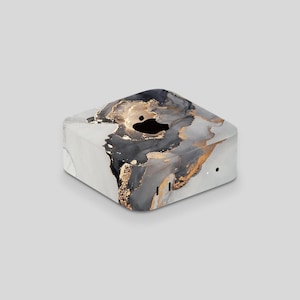 May include: A square device with a marble effect design. The surface has a mix of white, grey, black, and gold colours, forming an abstract pattern. The device has a black logo on top and small ports on the side.