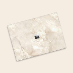 May include: A white marble laptop skin with a subtle beige pattern. The skin has a cutout for the Microsoft logo.