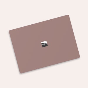 Könnte beinhalten: Ein geschlossener Laptop mit einem matten rosa oder mauvefarbenen Deckel. Das Microsoft Windows-Logo ist auf dem Deckel sichtbar.