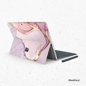 Könnte beinhalten: Eine rosa und goldene Marmor-Laptop-Haut mit einem goldenen Wirbelmuster. Die Haut ist auf einen geschlossenen Laptop mit einer schwarzen Tastatur und einem schwarzen Stift angebracht.