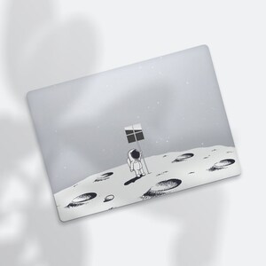May include: A laptop skin with a gray background and a black and white illustration of an astronaut standing on the moon holding a flag with the Windows logo. The moon has craters.