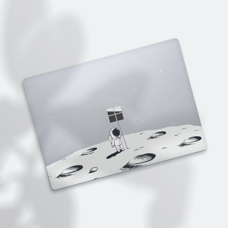 May include: A laptop skin with a gray background and a black and white illustration of an astronaut standing on the moon holding a flag with the Windows logo. The moon has craters.
