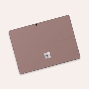Può includere: Un tablet Microsoft Surface color oro rosa con un obiettivo della fotocamera nero e un logo Windows argentato.