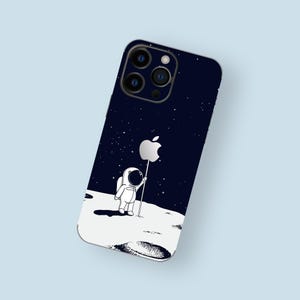 May include: A black phone case with a white astronaut holding an apple logo on a moon-like surface. The phone case has a starry night background.