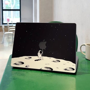 May include: A black laptop with a white astronaut holding an apple logo on the moon. The laptop is on a green surface.