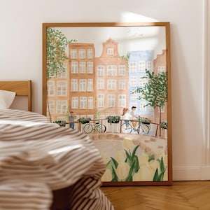 May include: A framed wall art print featuring a watercolour illustration of a European canal scene with tall, narrow buildings in shades of tan and blue, a stone bridge with people riding bicycles, and yellow tulips in the foreground.