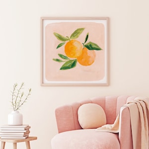 May include: A framed print of a painting of two orange citrus fruits with green leaves on a light pink background. The print is hanging on a white wall above a pink armchair with a throw blanket.