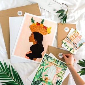 Puede incluir: Un conjunto de tres impresiones de arte que representan a una mujer con una cesta de fruta en la cabeza, una escena de jard&iacute;n tropical y una escena de calle de la ciudad. Las impresiones est&aacute;n en papel blanco con un fondo rosa claro y est&aacute;n empaquetadas en sobres marrones con pegatinas.