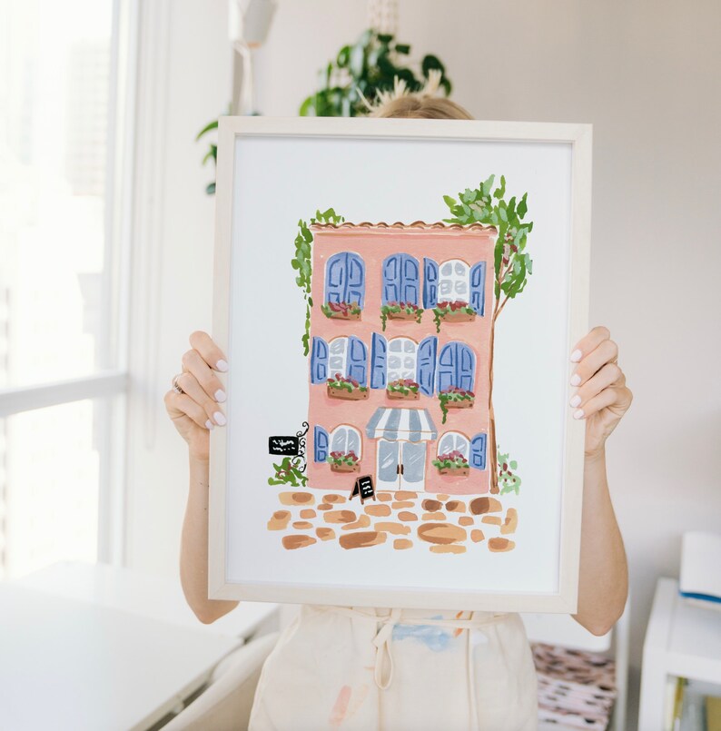 May include: A framed illustration of a pink building with blue shutters and a striped awning. The building has flowers in pots on the windowsills and a sign that says "Bakery".