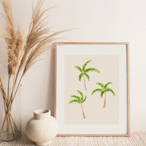 May include: A framed print featuring three watercolor palm trees with green fronds and reddish-brown trunks against a beige background. The artwork is displayed in a light wood frame, suggesting a tropical or coastal theme.