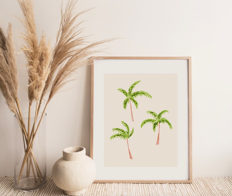 May include: A framed print featuring three watercolor palm trees with green fronds and reddish-brown trunks against a beige background. The artwork is displayed in a light wood frame, suggesting a tropical or coastal theme.