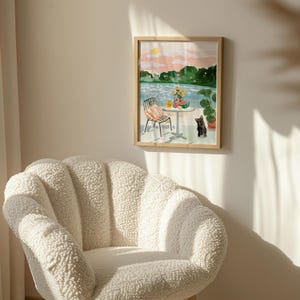 Puede incluir: Una pintura de acuarela de una escena de lago con una mesa, una silla y un gato negro. La pintura est&aacute; enmarcada en un marco de madera claro y cuelga de una pared beige claro.