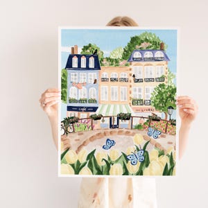 Op de afbeelding: Een aquarel schilderij van een Parijse straat sc&egrave;ne met kleurrijke gebouwen, een caf&eacute;, een boekwinkel en een brug met gele tulpen. De gebouwen hebben blauwe en groene accenten en er vliegen vlinders rond.