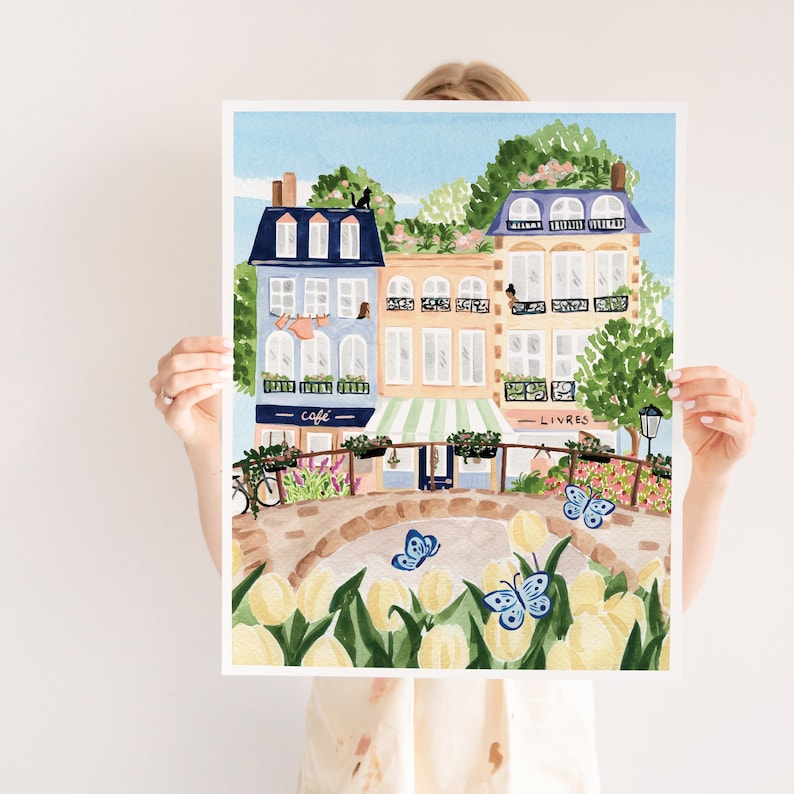 Op de afbeelding: Een aquarel schilderij van een Parijse straat sc&egrave;ne met kleurrijke gebouwen, een caf&eacute;, een boekwinkel en een brug met gele tulpen. De gebouwen hebben blauwe en groene accenten en er vliegen vlinders rond.