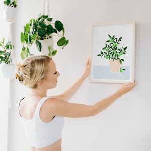 May include: A woman hangs a framed print of a potted lime tree with green leaves and fruit on a white wall. The print is in a light wood frame. The woman is wearing a white tank top and has blonde hair.