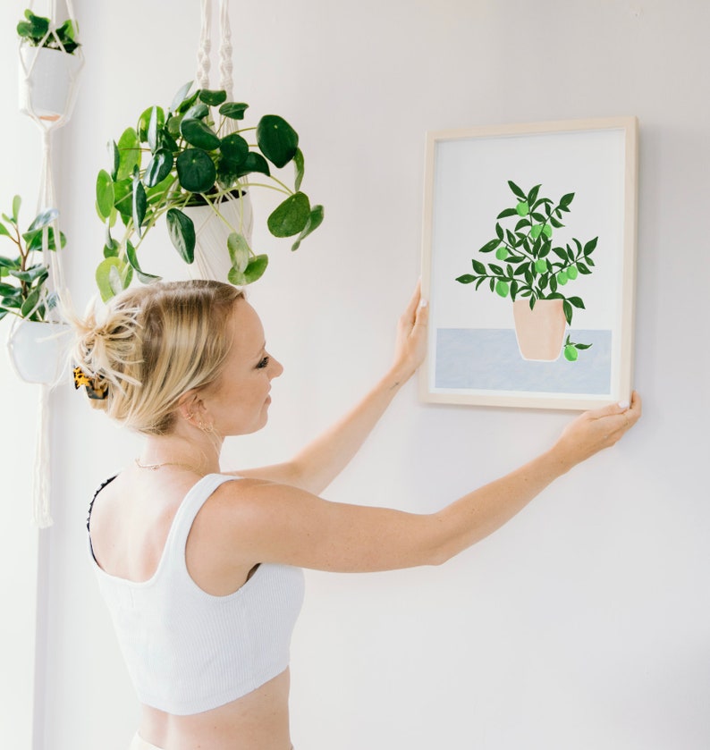 May include: A woman hangs a framed print of a potted lime tree with green leaves and fruit on a white wall. The print is in a light wood frame. The woman is wearing a white tank top and has blonde hair.