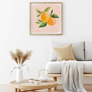 May include: A framed print of a painting of two orange citrus fruits with green leaves on a light pink background. The print is hanging on a white wall above a light brown couch with a white throw blanket and a woven coffee table with a white teapot and two white teacups.