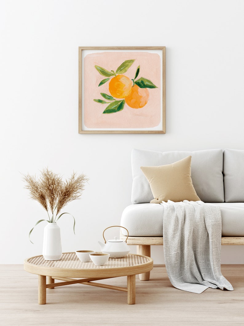 May include: A framed print of a painting of two orange citrus fruits with green leaves on a light pink background. The print is hanging on a white wall above a light brown couch with a white throw blanket and a woven coffee table with a white teapot and two white teacups.