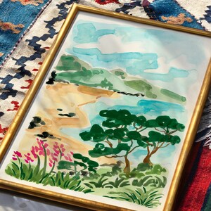 May include: A framed watercolor painting of a beach scene with a blue sky, white clouds, green hills, a sandy beach, and two green trees. The painting is in a gold frame.