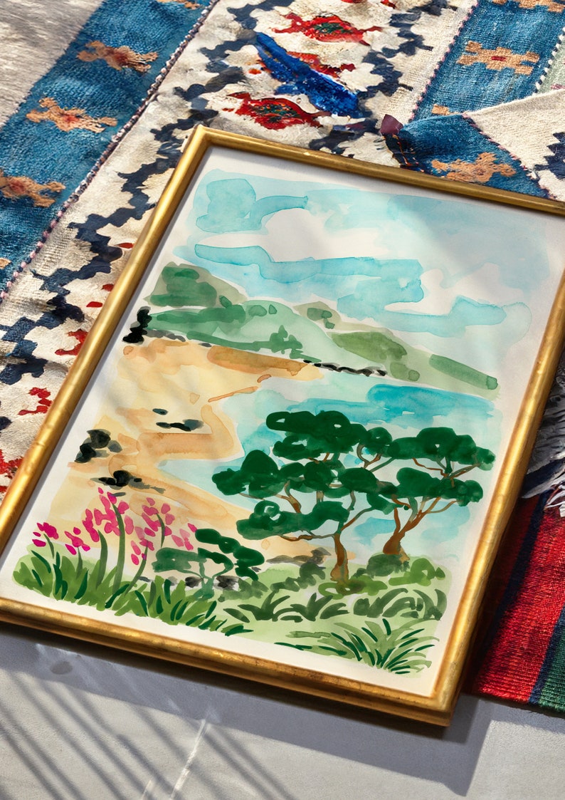 May include: A framed watercolor painting of a beach scene with a blue sky, white clouds, green hills, a sandy beach, and two green trees. The painting is in a gold frame.