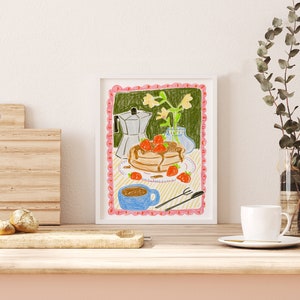 K&ouml;nnte beinhalten: Gerahmte Illustration mit einem Stapel Pfannkuchen mit Erdbeeren und Sirup, einer blauen Kaffeetasse und einer silbernen Espressomaschine. Das Kunstwerk hat einen rosa, wellenf&ouml;rmigen Rand und einen hellgr&uuml;nen Hintergrund. Auf einer Holzoberfl&auml;che, eine wei&szlig;e Tasse und Eukalyptus.