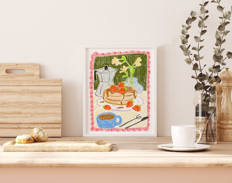 K&ouml;nnte beinhalten: Gerahmte Illustration mit einem Stapel Pfannkuchen mit Erdbeeren und Sirup, einer blauen Kaffeetasse und einer silbernen Espressomaschine. Das Kunstwerk hat einen rosa, wellenf&ouml;rmigen Rand und einen hellgr&uuml;nen Hintergrund. Auf einer Holzoberfl&auml;che, eine wei&szlig;e Tasse und Eukalyptus.