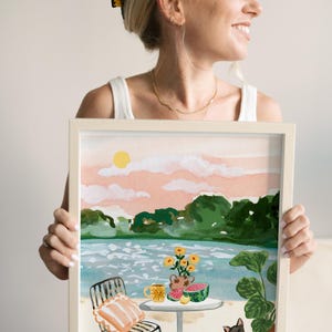Puede incluir: Una pintura al agua enmarcada que representa una escena de un lago con una mesa y una silla, un jarr&oacute;n de flores amarillas, una sand&iacute;a, limones y un perro negro. El cielo es rosa y naranja con un sol amarillo.
