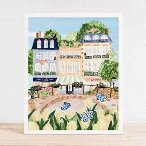 Op de afbeelding: Een aquarel schilderij van een Parijse straat sc&egrave;ne met kleurrijke gebouwen, een caf&eacute;, een boekwinkel en een brug met gele tulpen en blauwe vlinders. De tekst op het bord van het caf&eacute; luidt "caf&eacute;" en de tekst op het bord van de boekwinkel luidt "livres".