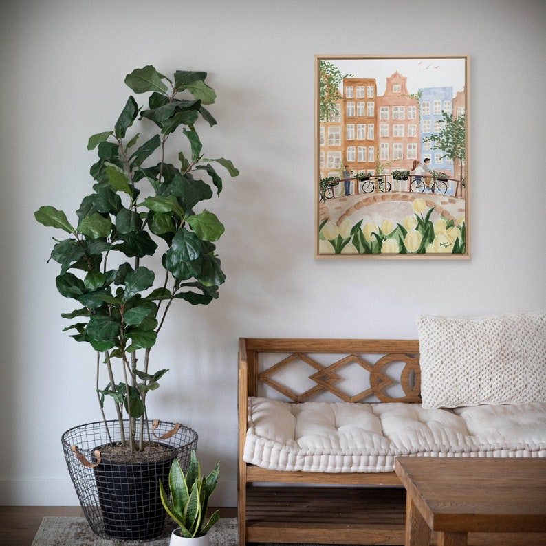 May include: A framed watercolour painting depicting a canal scene with tall, narrow buildings, a stone bridge, and people riding bicycles. The foreground features a field of pale yellow tulips. The artwork is displayed in a light wood frame.
