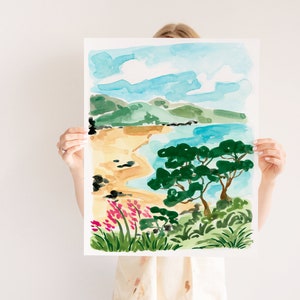 May include: A watercolor painting of a beach scene with a blue sky, green hills, a sandy beach, a blue ocean, and a green tree. The painting is framed in white.