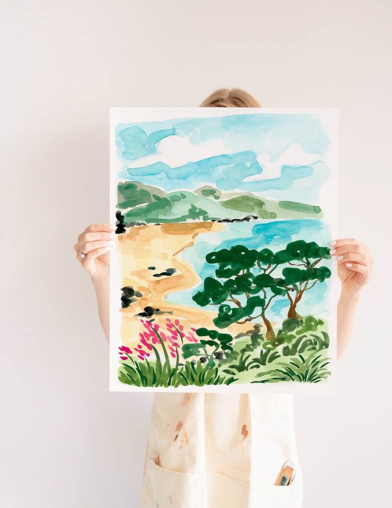 May include: A watercolor painting of a beach scene with a blue sky, green hills, a sandy beach, a blue ocean, and a green tree. The painting is framed in white.