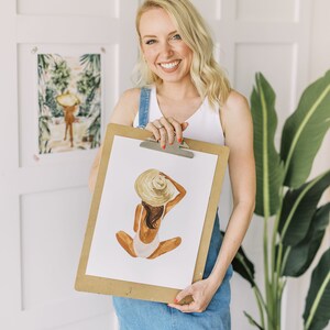 Op de afbeelding: Een vrouw houdt een klembord vast met een aquarelillustratie van een persoon die een witte badpak en een strohoed draagt. De illustratie is op een witte achtergrond.