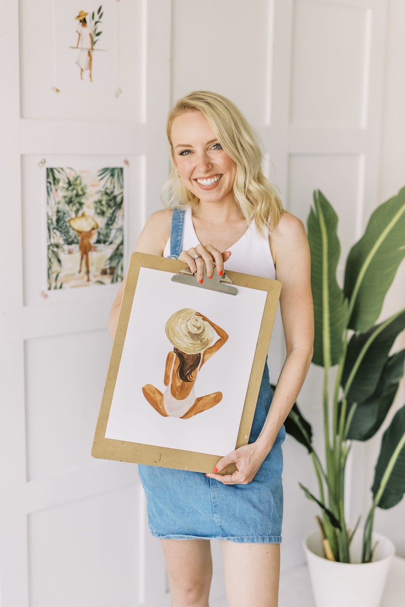 Op de afbeelding: Een vrouw houdt een klembord vast met een aquarelillustratie van een persoon die een witte badpak en een strohoed draagt. De illustratie is op een witte achtergrond.