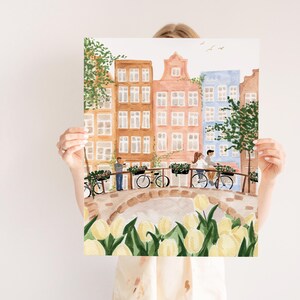 May include: A watercolour art print depicting a canal scene with tall, narrow buildings in shades of brown, peach, and blue. A stone bridge features people on bicycles and a person standing, with yellow tulips in the foreground and green trees.
